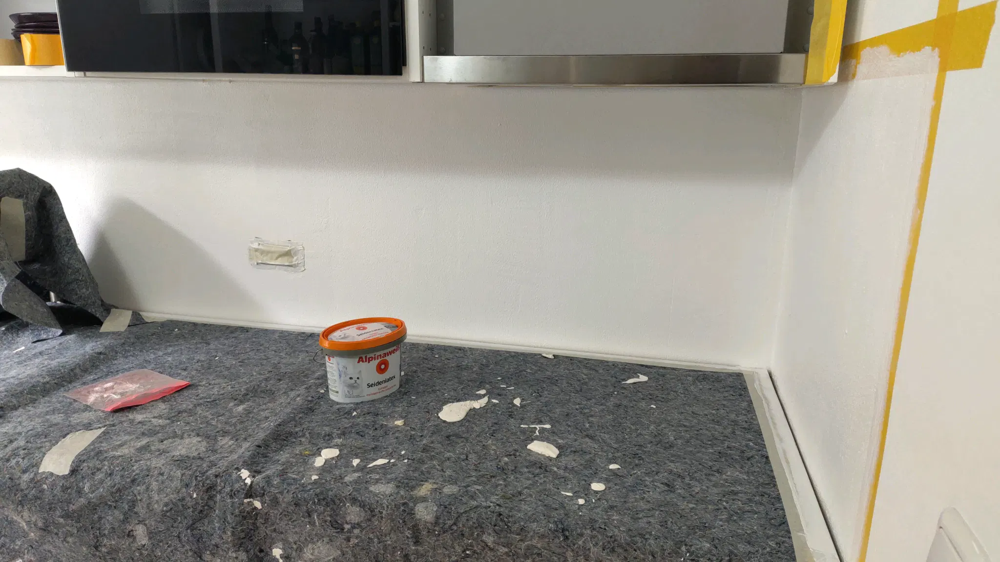 Covered kitchentop and yellow sticky tape on the wall. There's a small bucket of white latex paint. The tape is a bit white, indicating that the wall was painted. 